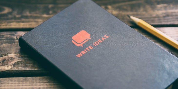 wooden table with pencil beside black journal with speech bubbles and the word "write ideas" on the cover