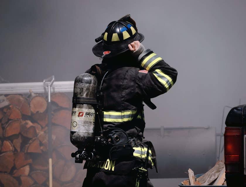 Ein Feuerwehrmann mit Brandschutzausrüstung steht vor einem Stapel Holz und neben einem Auto, umgeben von dickem Rauch.