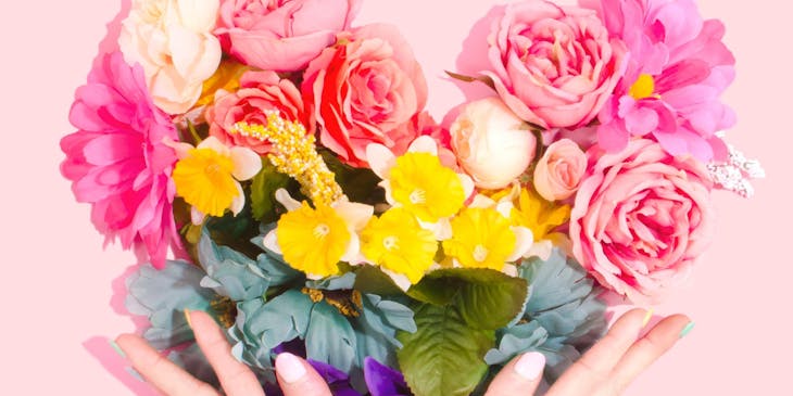 Hands holding a heart-shaped floral arrangement against a pink background.