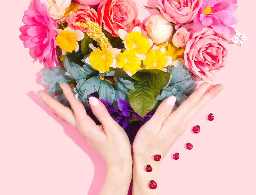Mãos segurando um arranjo floral em forma de coração contra um fundo rosa.