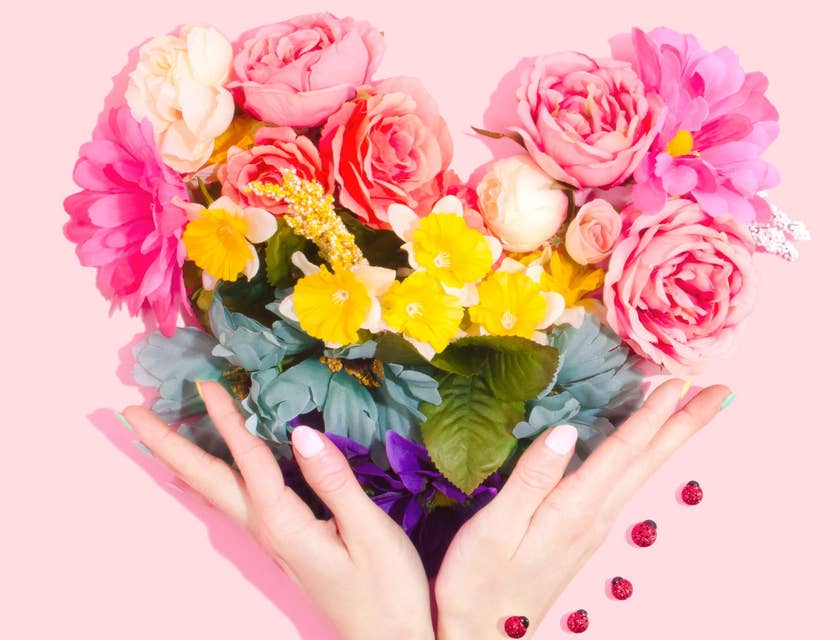 Des mains tenant une composition florale en forme de cœur sur un fond rose.