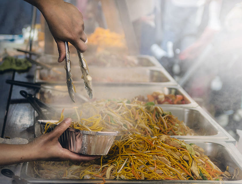 Person at a food stand dishing chow mein into a foil container.
