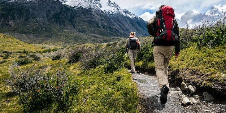 Two hikers walking a hiking trails towards a mountain covered in snow.