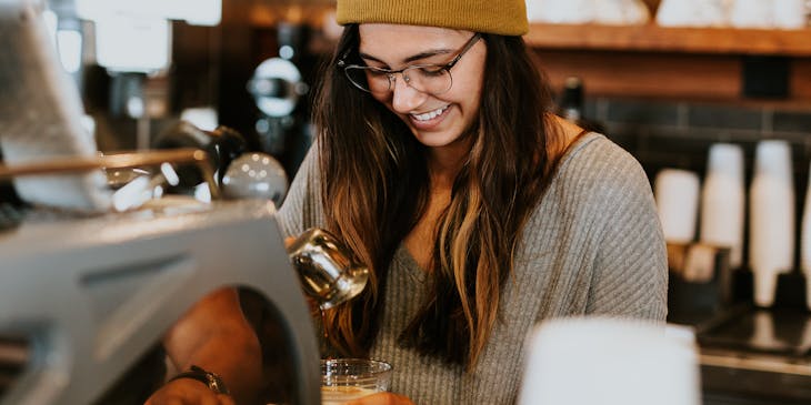 A hipster girl in beanie and glasses making coffee at a coffee shop.