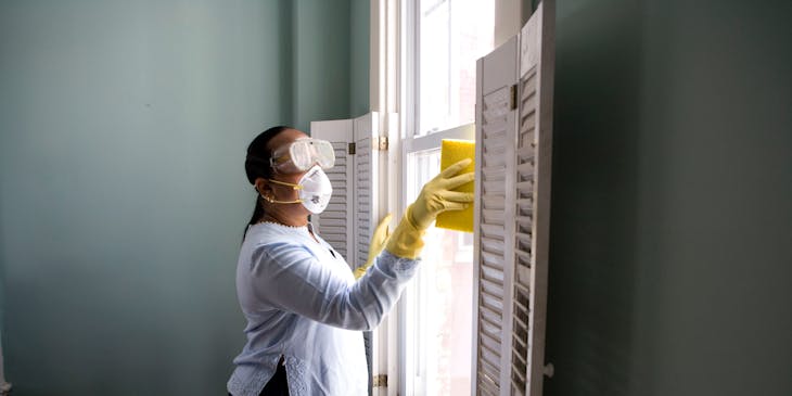 mujer con gafas y mascarilla limpiando una ventana con una esponja.