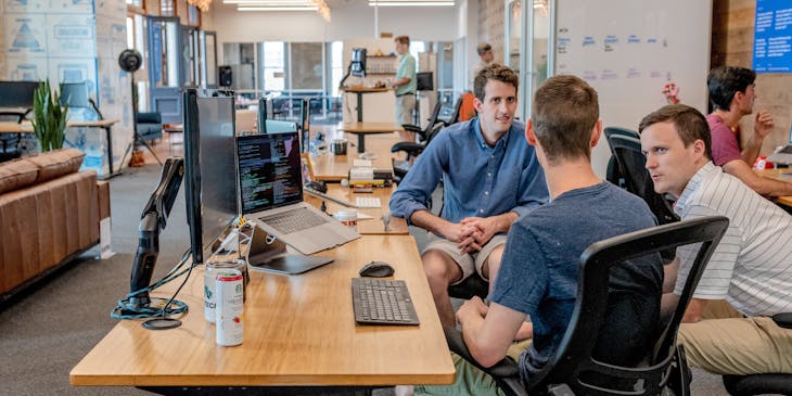 Three men talking around a laptop and monitor.