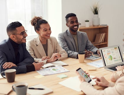 Group of three people sitting at a desk facing a woman with a cellphone in her hand