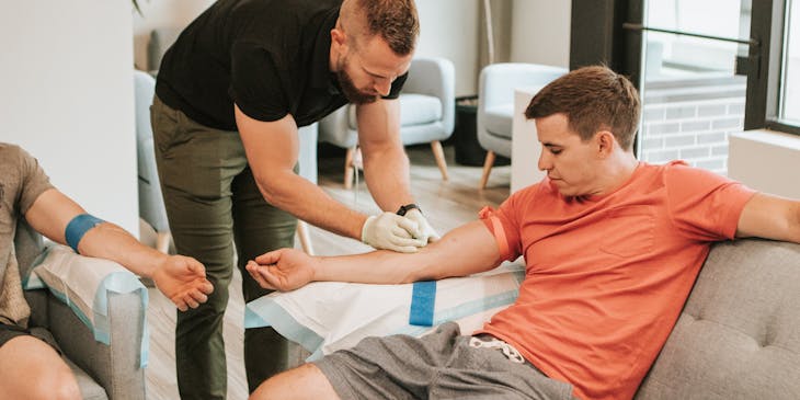 Male nurse inserting IV lines secured with blue tape into the arms of two male patients.