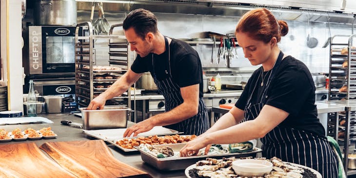 Male and female chefs wearing dark shirts and striped aprons while preparing food in a kitchen.