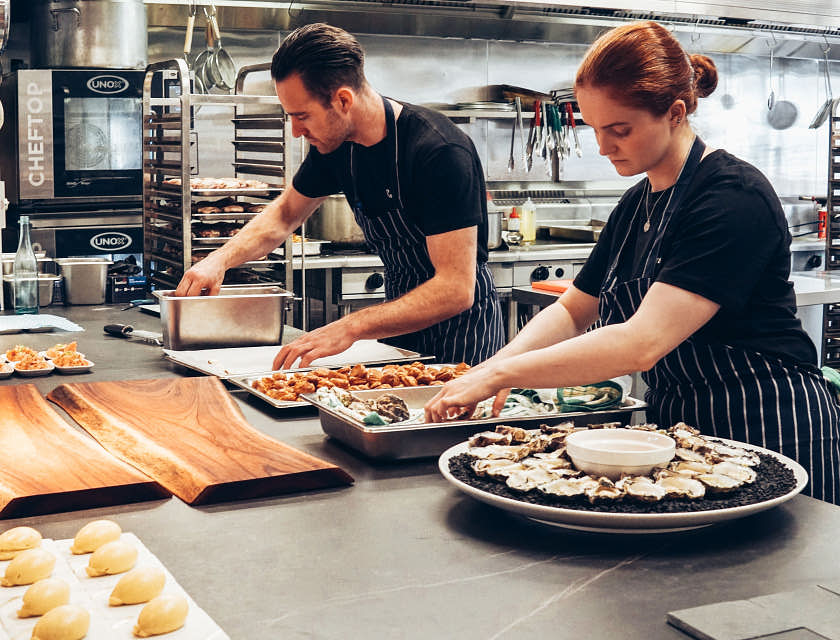 Male and female chefs wearing dark shirts and striped aprons while preparing food in a kitchen.