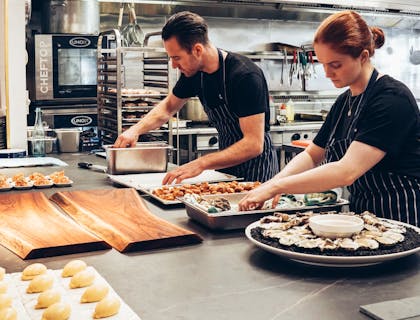 Des cuisiniers et des cuisinières portant des chemises sombres et des tabliers rayés alors qu'ils préparent des aliments dans une cuisine partagée.