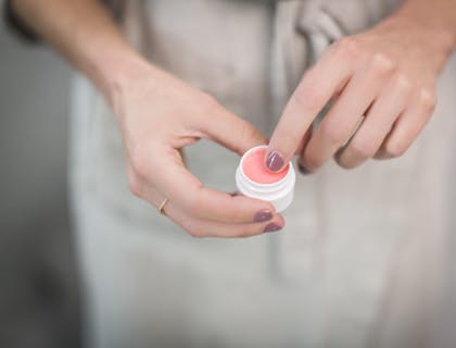 Un gros plan des mains d'une femme tenant un petit récipient rempli de baume à lèvres rose.