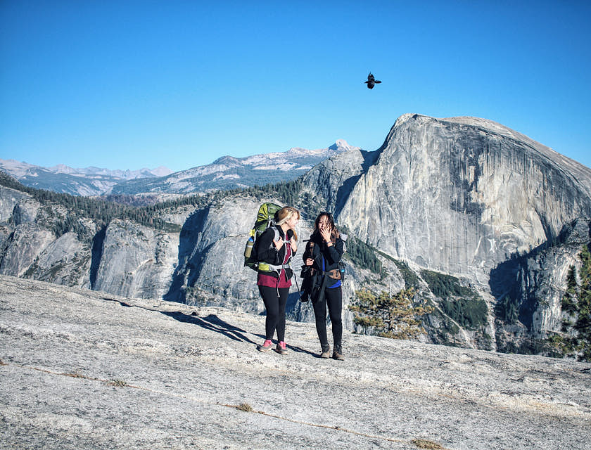 Dos mujeres con mochilas en la espalda caminando en una montaña y un ave volando al fondo en una empresa de turismo de aventura.