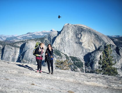 Dos mujeres con mochilas en la espalda caminando en una montaña y un ave volando al fondo en una empresa de turismo de aventura.