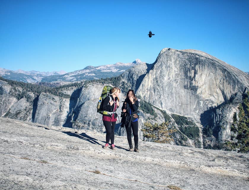 Dos mujeres con mochilas en la espalda caminando en una montaña y un ave volando al fondo en una empresa de turismo de aventura.