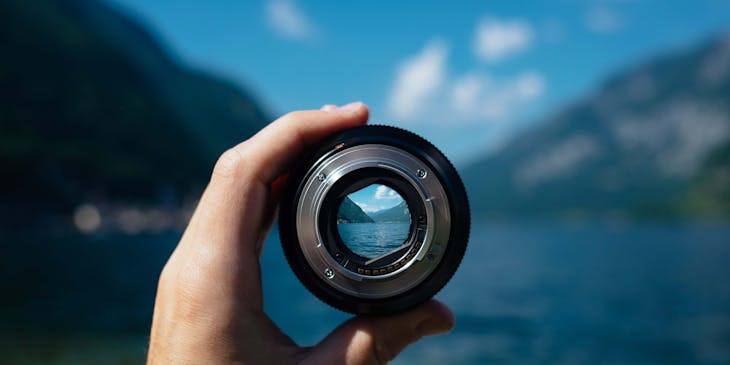 A nature photographer holding a camera lens up to the mountain lake view in the background.