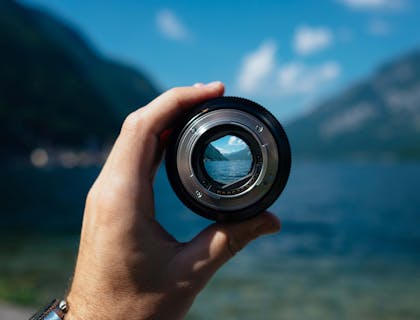 Un fotografo naturalistico che tiene in mano un obiettivo fotografico davanti a un paesaggio montano che si intravede sullo sfondo.