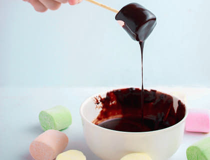 Una persona sosteniendo un bombón con un palillo mientras lo cubre de chocolate deterrido en una bombonería.