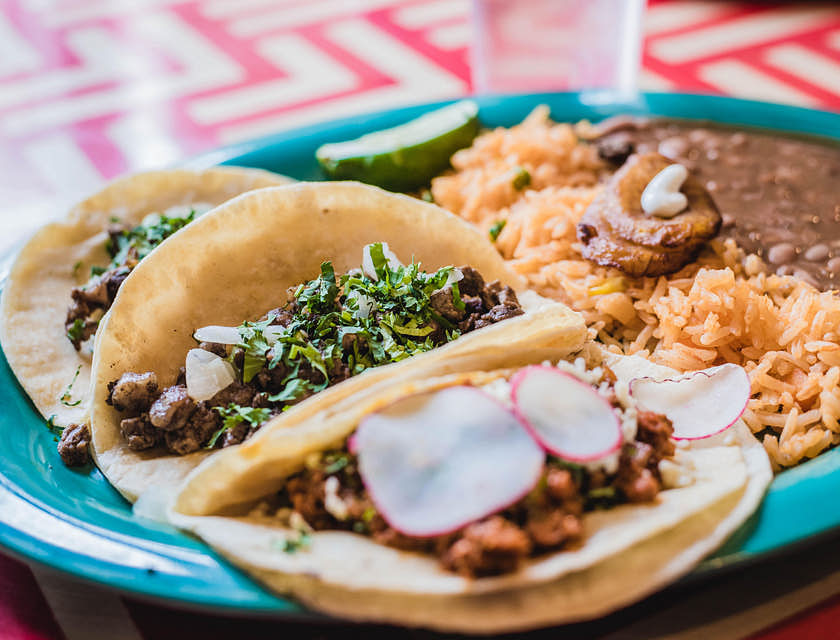 Un plato con tres tacos, frijoles refritos y arroz sobre una mesa en una cenaduría.