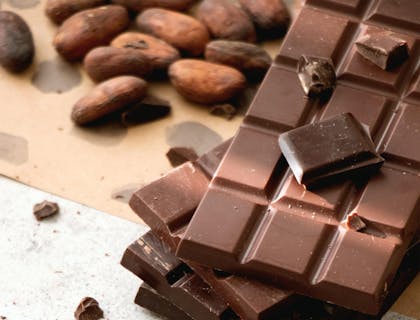 Cuatro barras de chocolate encimadas y trozos de chocolate esparcidos con semillas de cacao Amazonas al fondo sobre un mantel en una mesa de granito.