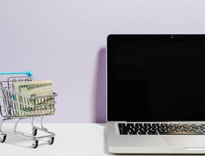 La imagen de un carrito de compras miniatura con billetes dentro frente a una laptop en una empresa de e-commerce.