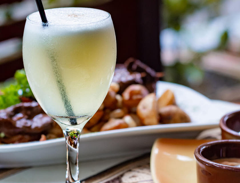 Bebida a base de pisco sobre una mesa y alimentos en un plato al fondo.