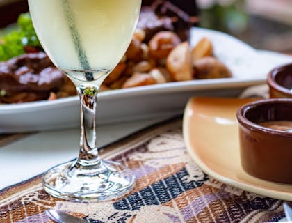 Bebida a base de pisco sobre una mesa y alimentos en un plato al fondo.