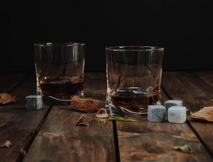 Dos vasos servidos con ron de una empresa de ron venezolano sobre madera con hojas secas esparcidas y cubos de azúcar.
