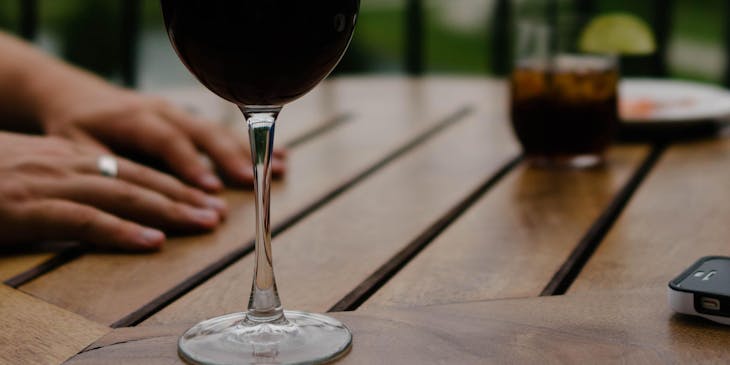 Una copa con vino de una empresa de vino de malbec sobre una mesa de madera con unas manos de mujer un vaso con refresco y un barandal con luces encendidas al fondo.