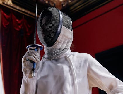 Un esgrimista con careta y la espada frente a su cara con una pared ventana y cortinas al fondo en una escuela de esgrima.