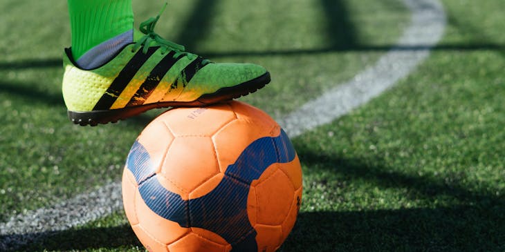 Un jugador de fútbol usando zapatillas y calcetas amarillas con su pie sobre un balón anaranjado en una cancha de una escuela de fútbol.