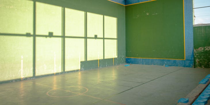 Una cancha en una escuela de pelota vasca con el reflejo del sol en una pared y techo de lamina corrugada.
