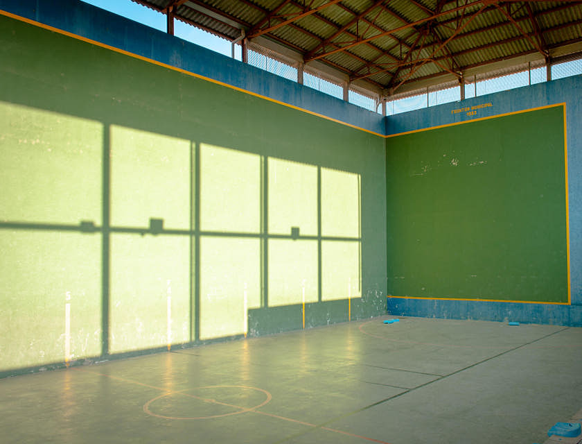Una cancha en una escuela de pelota vasca con el reflejo del sol en una pared y techo de lamina corrugada.
