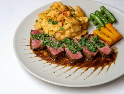 Un corte de carne a termino medio y mac and cheese con chimichurri encima servido en un plato.