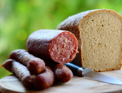Unos trozos de chorizo a un lado de una pieza de pan blanco y un cuchillo sobre una tabla de madera para picar fuera de un negocio de choripán.