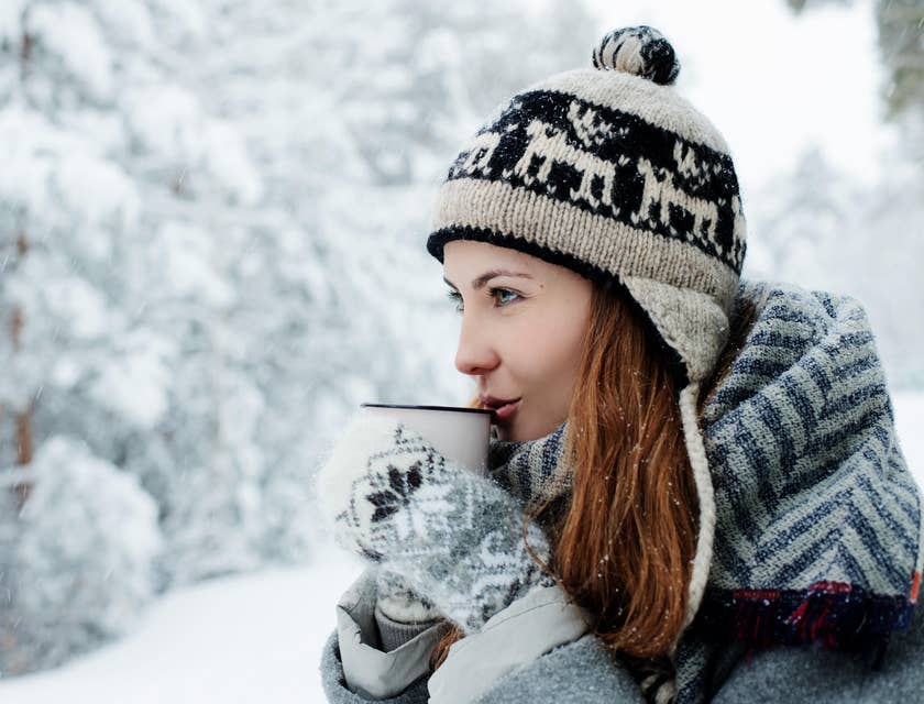 Una mujer en un bosque nevado tomando café usando un gorro de un negocio de chullos.