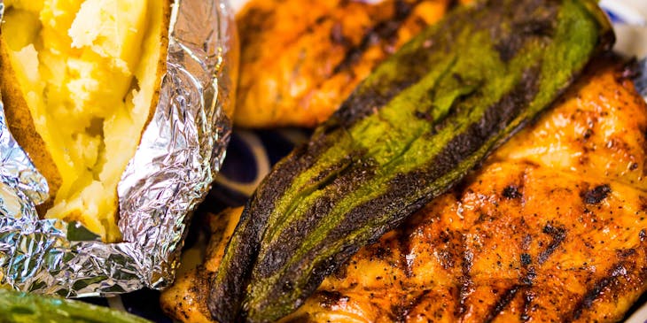 Una papa asada con un par de pechugas de pollo asadas y chiles toreados sobre un plato blanco con detalles azules.