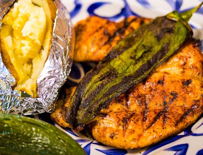 Una papa asada con un par de pechugas de pollo asadas y chiles toreados sobre un plato blanco con detalles azules.