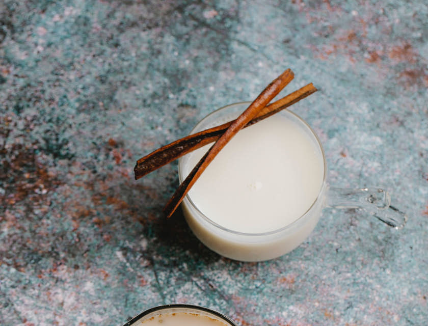 Dos vasos con coquito, canela sin procesar y un pedazo de chocolate, sobre una mesa en un negocio de coquito.