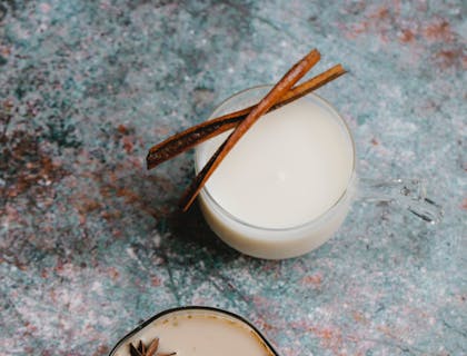 Dos vasos con coquito, canela sin procesar y un pedazo de chocolate, sobre una mesa en un negocio de coquito.