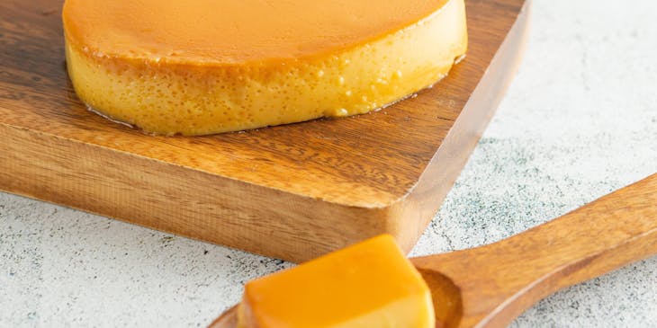 Un dulce de batata sin cortar sobre una tabla de madera junto a una cuchara de madera y una canasta de huevos en un negocio de dulces de batata.