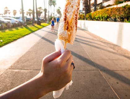 Persona sujetando un elote de un negocio de elote loco en un camino rodeado por palmeras y un edificio al lado.