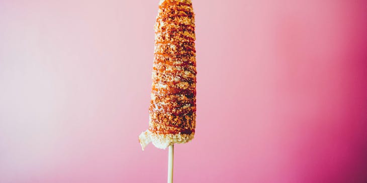 Un elote con mayonesa y chile en polvo con queso rallado espolvoreado sobre un fondo color rosa.