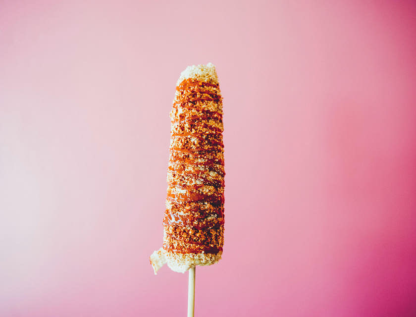un elote con mayonesa y chile en polvo con queso rallado espolvoreado sobre un fondo color rosa