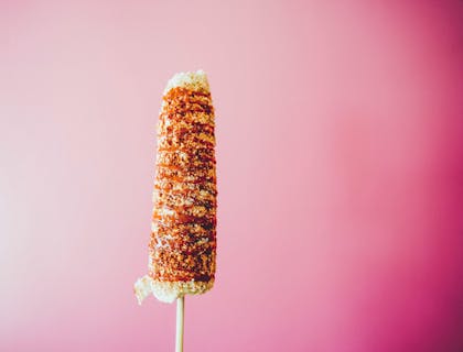 Un elote con mayonesa y chile en polvo con queso rallado espolvoreado sobre un fondo color rosa.