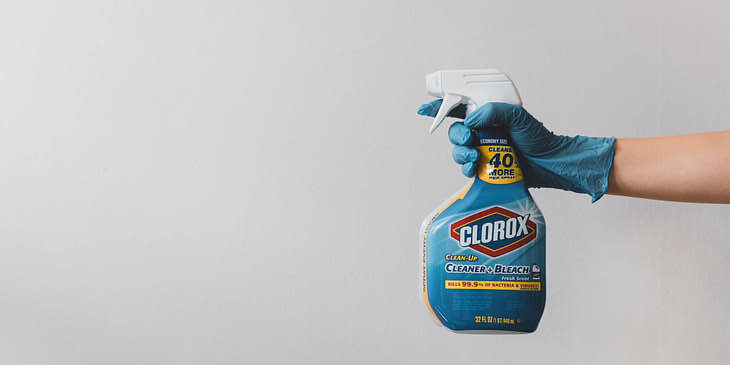 Una mano con un guante quirúrgico azul sosteniendo una botella de cloro a punto de esterilizar y desinfectar un espacio.