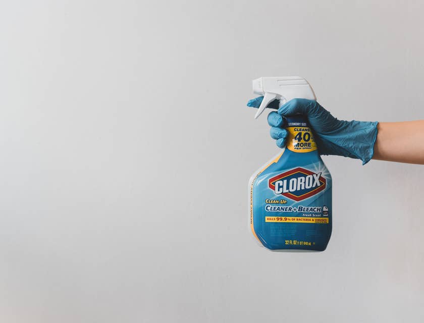 Una mano con un guante quirúrgico azul sosteniendo una botella de cloro a punto de esterilizar y desinfectar un espacio.