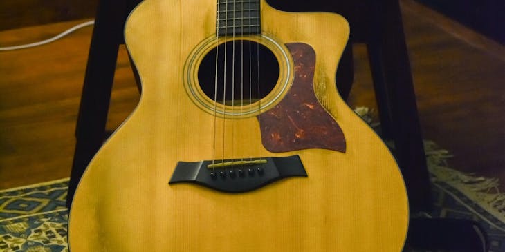 Una guitarra acústica de color café recargada en una silla de madera sobre un tapete en un suelo de madera con un fondo de pared azul oscuro en un negocio de guitarras.