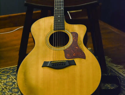 Una guitarra acústica de color café recargada en una silla de madera sobre un tapete en un suelo de madera con un fondo de pared azul oscuro en un negocio de guitarras.