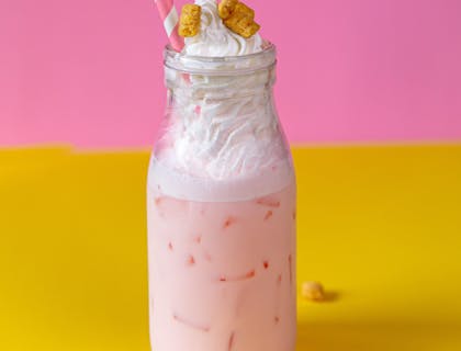 Una malteada de fresa con crema batida encima y trozos de galleta frente a un fondo rosa y amarillo en un negocio de malteadas.